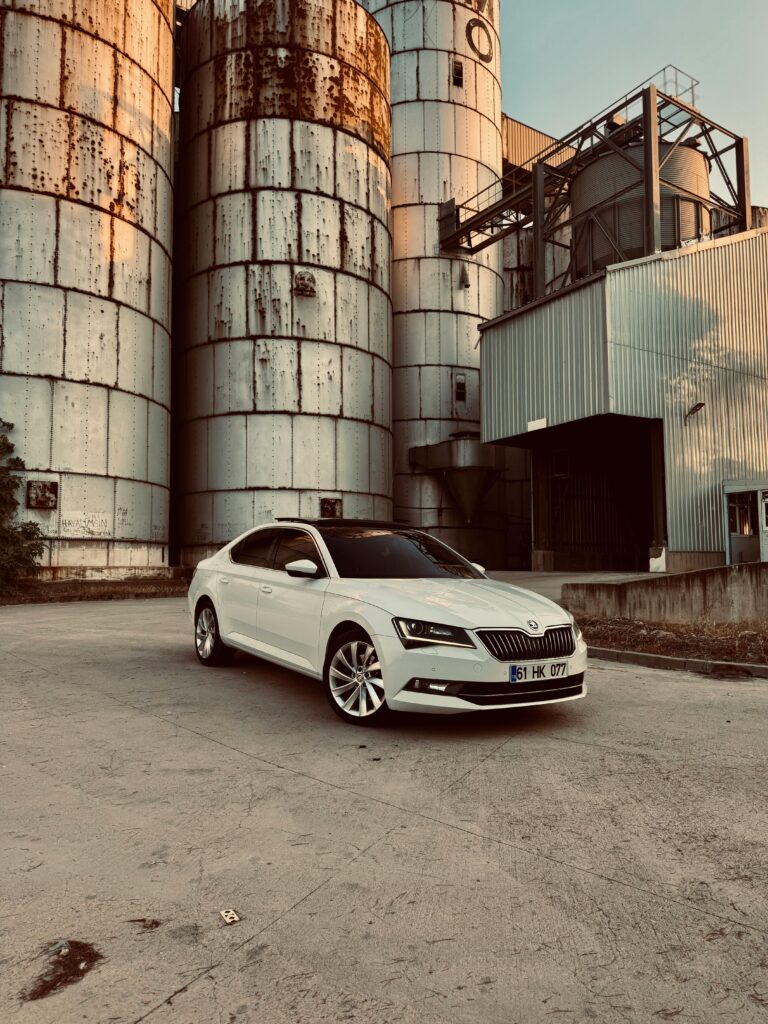 A white Skoda parked in front of abandoned industrial silos under a sunny sky.